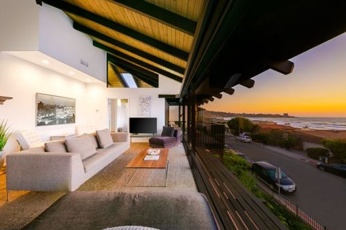 Modern living room with a coastal view at sunset, featuring large windows, a sofa, and stylish decor.