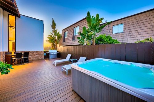 A serene outdoor space featuring a hot tub, lounge chairs, and a grill, surrounded by greenery and modern buildings.