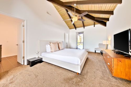 Bright bedroom with a high ceiling, featuring a white bed, wooden furniture, and a large window.