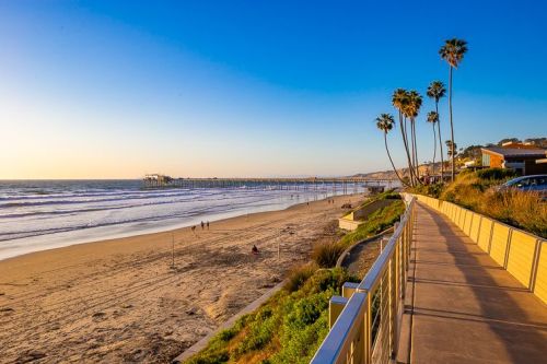A scenic beach view at sunset, featuring a walkway, palm trees, and a pier in the distance.