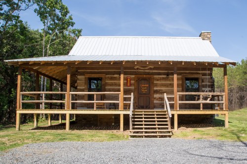 A rustic log cabin with a metal roof, wooden porch, and steps, surrounded by trees and gravel.