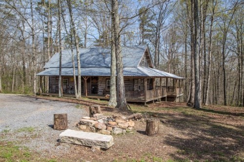 A rustic log cabin surrounded by trees, featuring a porch and a stone fire pit in the foreground.
