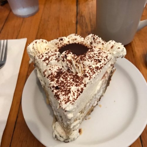 A slice of creamy cake topped with whipped cream and chocolate shavings, served on a white plate.