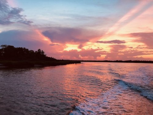 A serene river at sunset, reflecting vibrant pink and orange hues in the sky, with silhouettes of trees along the shore.