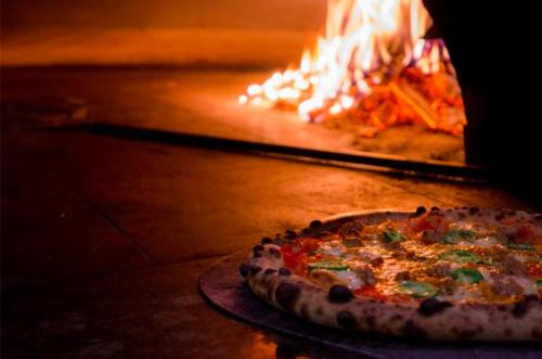 A freshly baked pizza sits on a stone surface, with a wood-fired oven and flames in the background.