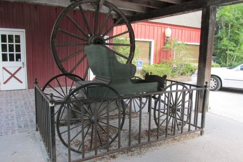 An old-fashioned horse-drawn carriage displayed in a fenced area under a wooden structure.