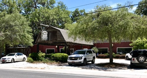 A rustic building with a wooden sign, surrounded by trees and parked cars on a sunny day.