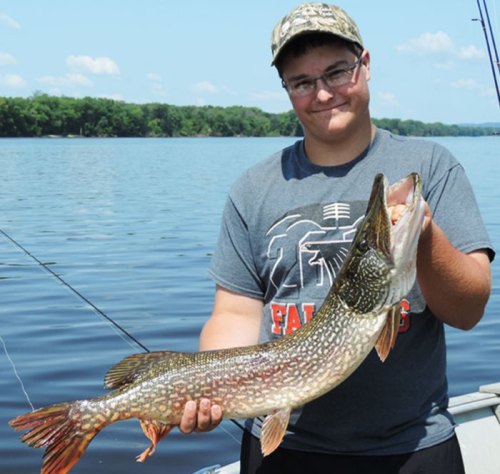 A young man in a cap holds a large pike fish while standing on a boat, with a calm lake and trees in the background.