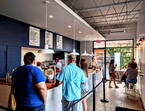 A modern café interior with customers in line, a menu board, and people seated at tables in the background.