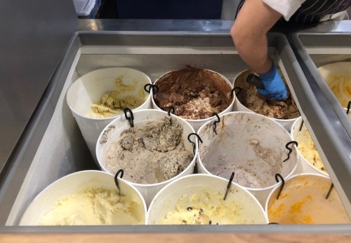 A variety of ice cream flavors in large containers, with a person scooping from one of the tubs.