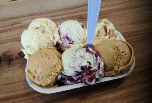 A dish of six ice cream scoops in various flavors, with a white spoon resting on top, on a wooden surface.