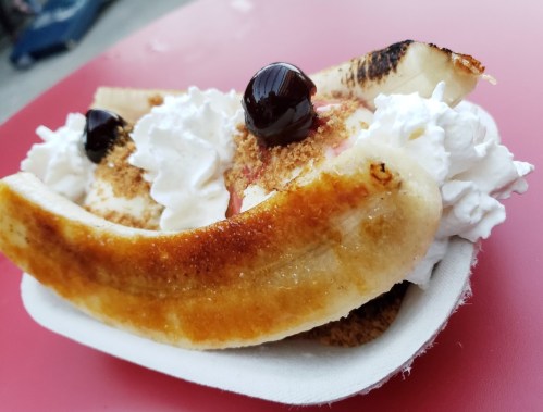 A banana split topped with whipped cream and cherries, served in a paper boat on a red table.