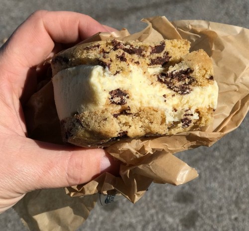 A hand holding a half-eaten cookie sandwich filled with vanilla ice cream and chocolate chunks, wrapped in brown paper.