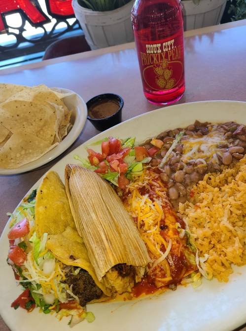A plate of tacos, tamale, rice, beans, and fresh toppings, with a side of chips and a bottle of prickly pear soda.