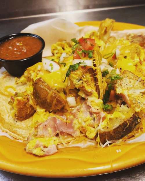 A plate of nachos topped with cheese, ham, scrambled eggs, onions, and tomatoes, served with a side of salsa.