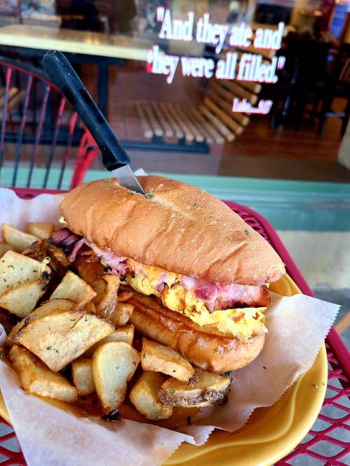 A sandwich with ham and egg, topped with a knife, served with golden-brown potato wedges on a red tray.