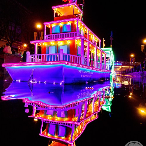 A brightly lit boat with colorful lights reflecting on the water at night, surrounded by festive decorations.