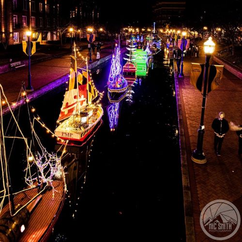 Colorful boats adorned with lights float along a canal at night, surrounded by festive decorations and a brick walkway.