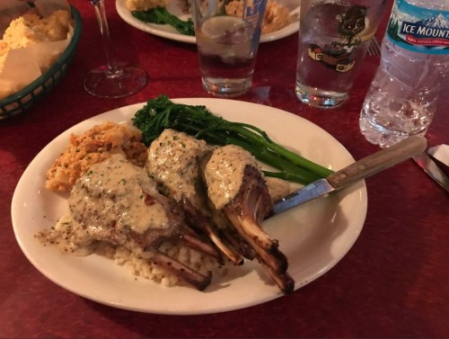 A plate of lamb chops with creamy sauce, served with rice and broccolini, alongside glasses of water.