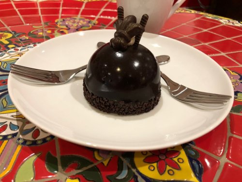 A glossy chocolate dome dessert on a white plate, garnished with chocolate curls, served with two forks.