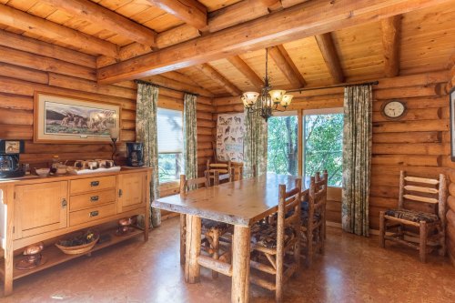 Cozy log cabin dining area with a large wooden table, chairs, and natural light from windows. Rustic decor and warm tones.