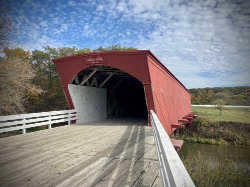 A red covered bridge spans a river, surrounded by autumn trees and a cloudy sky.