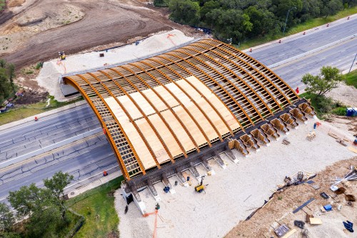 Aerial view of a large, curved wooden structure under construction beside a road, with surrounding earthworks and materials.
