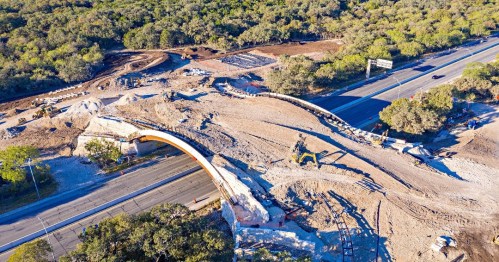 Aerial view of a construction site with a partially built bridge over a road, surrounded by trees and machinery.