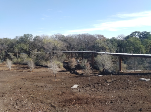 A long, elevated walkway stretches over a barren landscape with sparse trees and clear blue skies in the background.