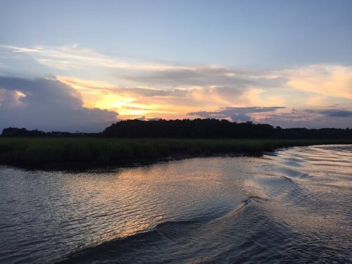A serene sunset over a calm river, with reflections on the water and lush greenery along the shore.