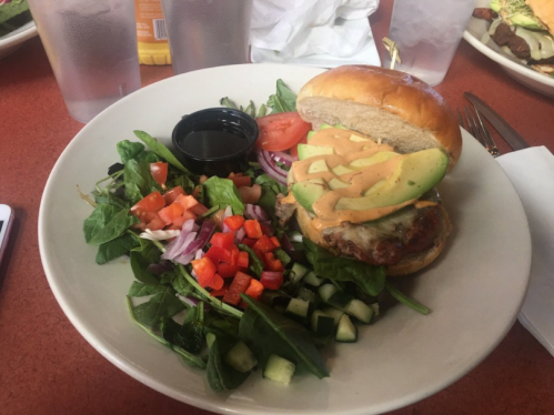 A plate with a burger topped with avocado and sauce, served with a fresh salad of mixed greens and diced vegetables.