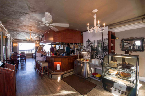 Cozy café interior with a bar, chandelier, and display case of desserts, featuring wooden furniture and a beach view.
