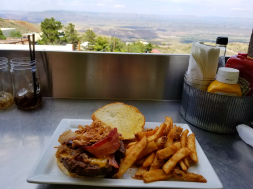 A plate with a burger topped with bacon, crispy onions, and fries, set against a scenic mountain view.
