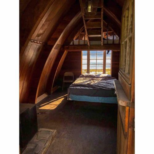 Cozy wooden cabin interior with a bed, chair, and large windows overlooking a scenic view.