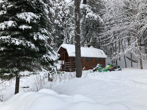 A cozy log cabin surrounded by snow-covered trees, with snowmobiles parked nearby in a winter landscape.
