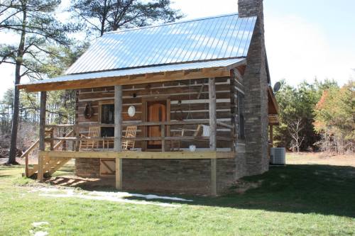 A rustic log cabin with a stone foundation, wooden porch, and rocking chairs, surrounded by trees and grass.