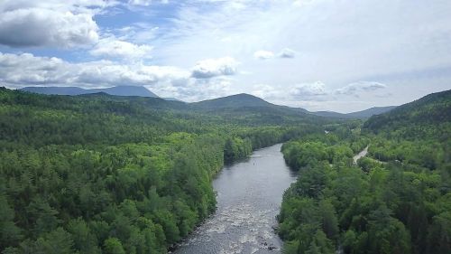 Aerial view of a winding river surrounded by lush green forests and mountains under a partly cloudy sky.
