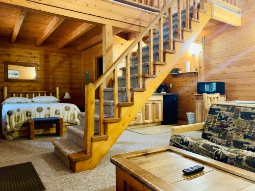 Cozy wooden cabin interior with a staircase, bed, sofa, and kitchenette, featuring warm lighting and rustic decor.