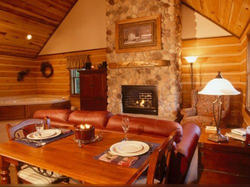 Cozy cabin interior with a stone fireplace, wooden walls, dining table set for two, and a comfortable armchair.