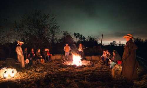 A group of people gathered around a campfire at night, sitting on hay bales, with a dark sky and trees in the background.