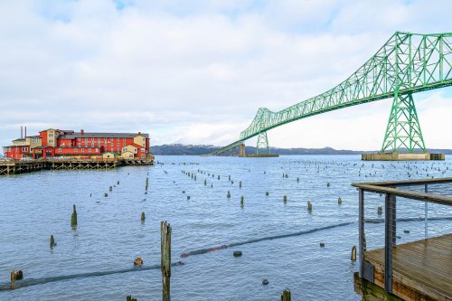A green bridge spans a river, with a red building on the shore and wooden posts emerging from the water.