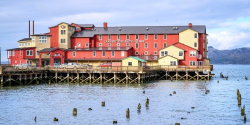 A red and yellow hotel on a pier, surrounded by water and wooden posts, under a cloudy sky.