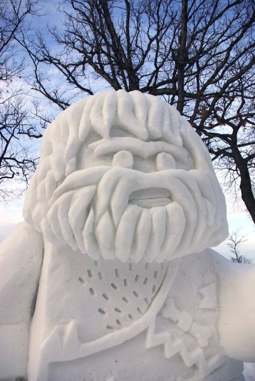 A large, detailed snow sculpture of a bearded figure with a rugged appearance, set against a winter sky and bare trees.