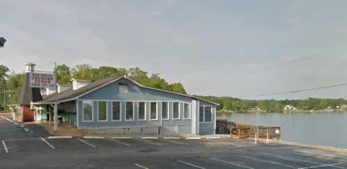 A lakeside restaurant with large windows, surrounded by trees and a parking lot, under a clear blue sky.