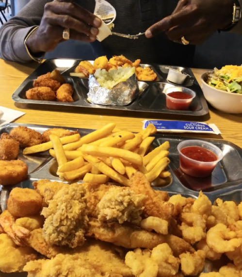 A table with two trays of fried seafood, fries, and sides, with a person preparing food in the background.