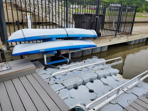Two kayaks on a rack above a floating dock, with a wooden walkway and water in the background.