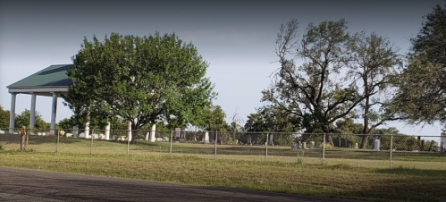 A fenced cemetery with trees and a gazebo, set against a clear sky.