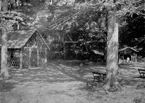 Black and white image of a wooded area with two rustic structures and benches, surrounded by trees.
