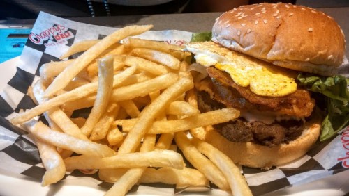 A sesame seed bun burger with lettuce, a fried egg, and crispy hash browns, served with a side of French fries.