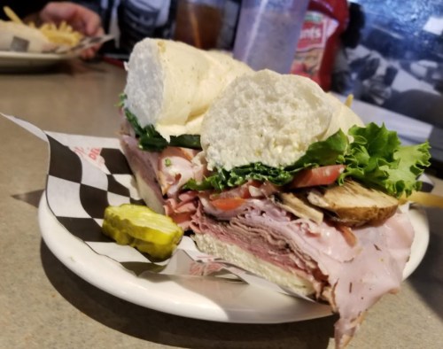 A hearty sandwich with layers of ham, lettuce, tomato, and mushrooms, served with a pickle on the side.